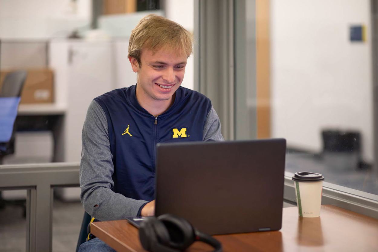 A smiling person sits at a table using a laptop in a bright office space. They are wearing a navy and gray University of Michigan zip-up top with a yellow "M" logo, and a takeaway coffee cup and a pair of headphones rest on the table beside the computer.