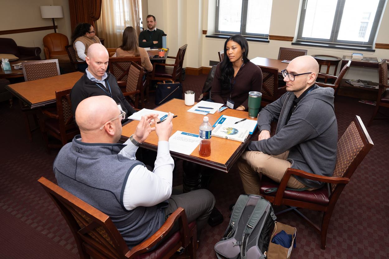 A group of four people sit at a table having a discussion, with worksheets and beverages in front of them.