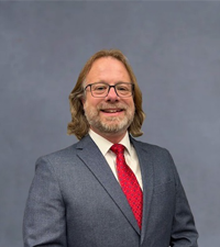 A smiling man with shoulder-length light brown hair, a beard, and glasses wears a blue suit and red tie.