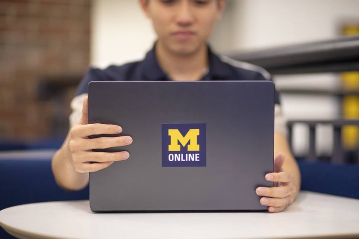 A person sits behind a laptop displaying the University of Michigan “M Online” logo.