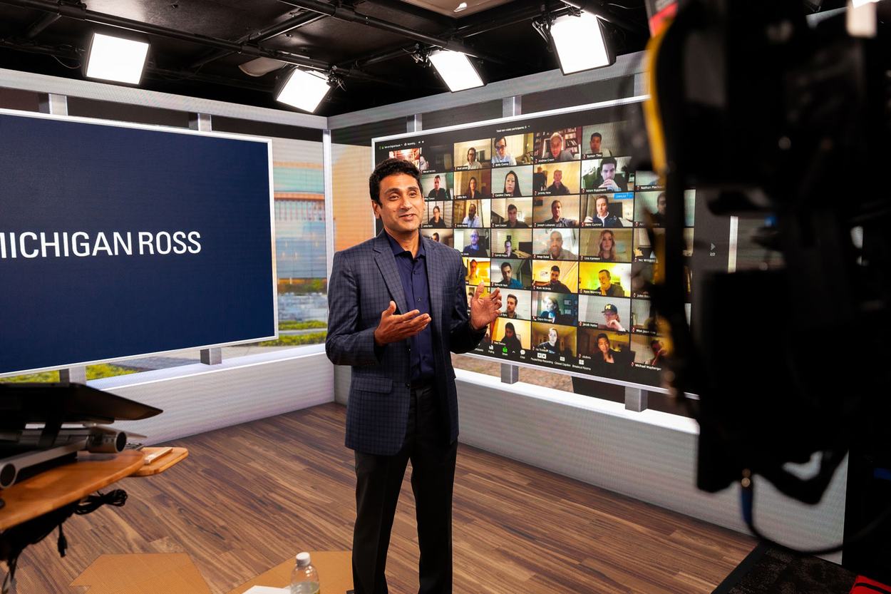 A man wearing a dark suit records a lecture in a studio with Michigan Ross on a background screen.
