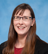 A smiling woman with long brown hair and glasses wears a black blazer over a red top against a blue background.