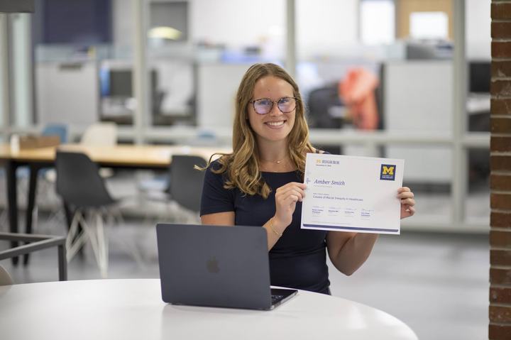 A student sits at a table with a laptop holding up a Michigan Online certificate.