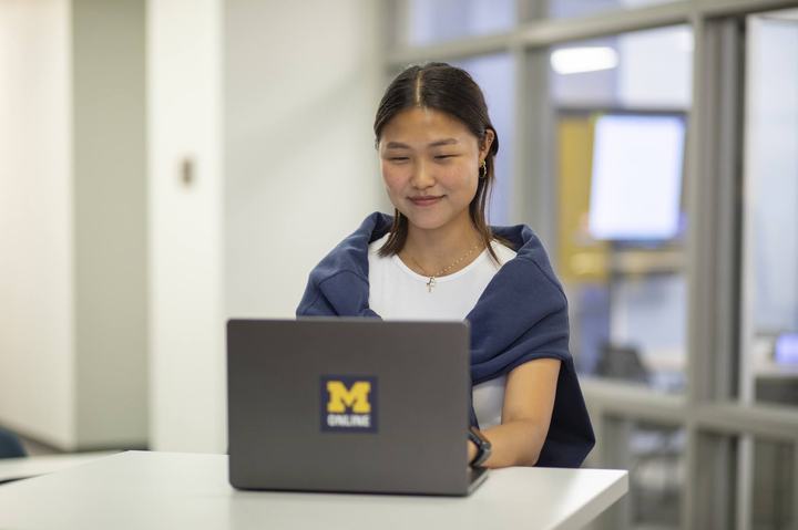 A U-M student sits at a table working on a laptop with an “M Online” logo on the back.