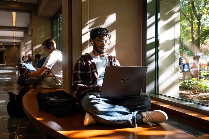 Male undergraduate student typing on a laptop next to a window